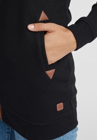 Svart sweatshirt med en lomme med en brun trekantet aksent og en firkantet lærmerke på den nedre siden. Glatt, teksturert stoff.