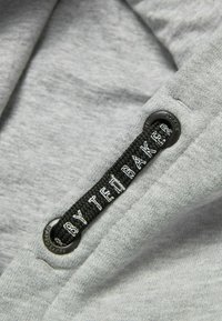 Grijze stof met een textuur, voorzien van een zwart geweven label waarop "BY TED BAKER" staat, bevestigd met een metalen oog.