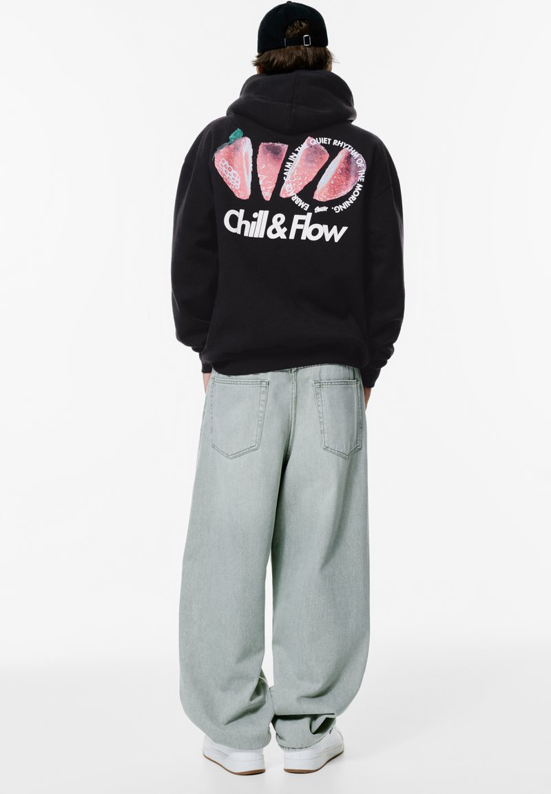 Hanorac negru cu un grafic mare de căpșună și textul "Chill & Flow" pe spate; asortat cu blugi oversized de culoare verde deschis și adidași albi.