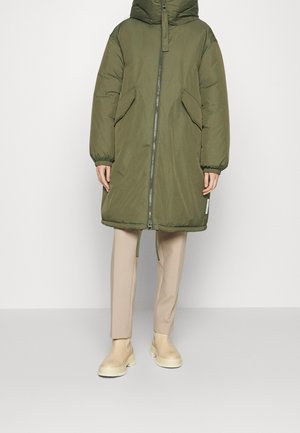 Person trägt langen olivgrünen gesteppte Mantel mit Frontreißverschluss, schrägen Klappentaschen, beige Hose und cremefarbene Knöchelstiefel vor einfarbigem Hintergrund.