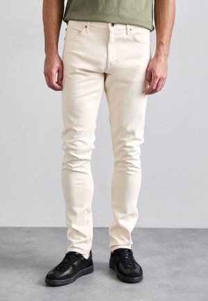 Hombre con jeans ajustados blanco roto y zapatillas negras, de pie sobre un suelo gris frente a una pared gris claro.