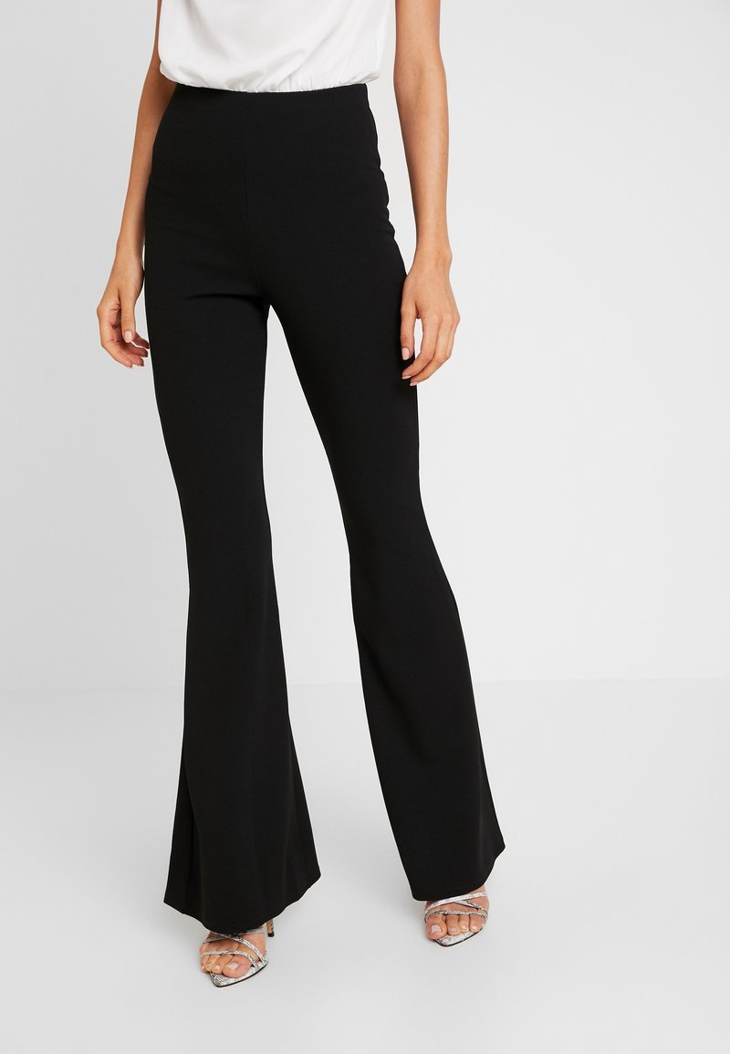 Femme portant un pantalon noir taille haute évasé et des talons argentés à brides, associé à un haut blanc rentré, debout devant un fond uni.