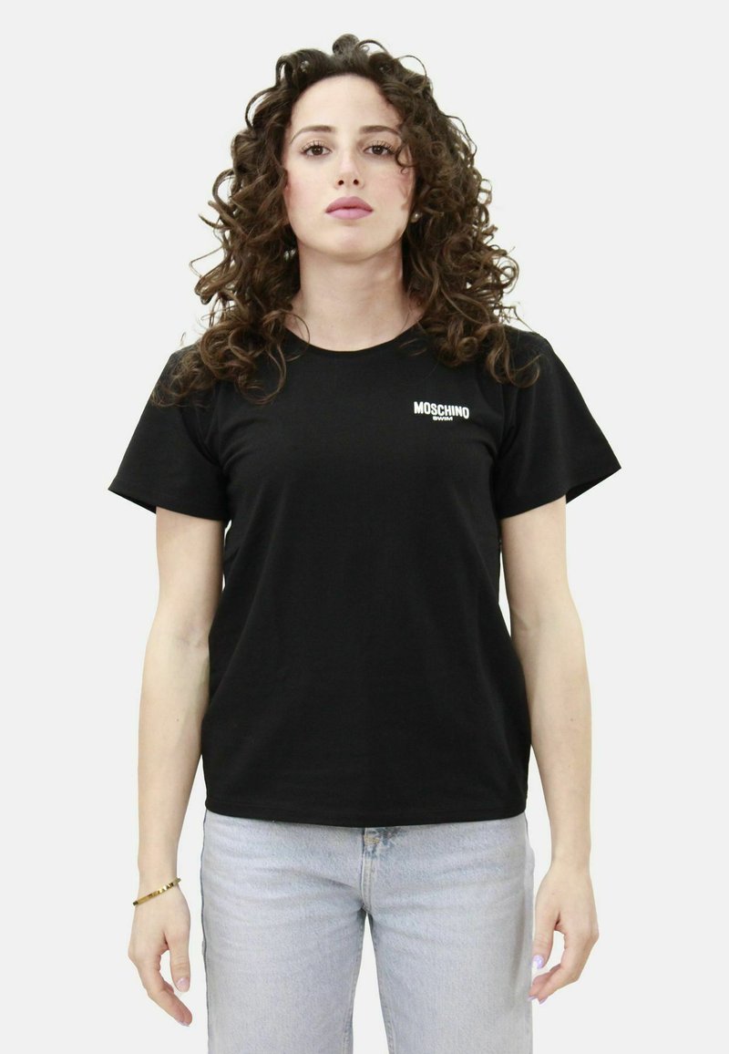 Maglietta in cotone nera con maniche corte e scollo rotondo, caratterizzata da un logo Moschino bianco sul lato sinistro del petto. Vestibilità casual, design semplice.