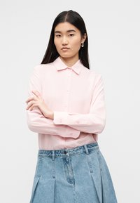 Ανοιχτό ροζ μπλούζα με μακριά μανίκια και γιακά, με κουμπιά στο μπροστινό μέρος και μανσέτες, συνδυασμένη με φούστα από τζιν.