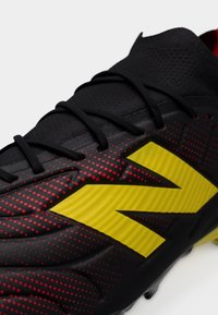 Μαύρο ποδοσφαιρικό παπούτσι με υφή στην επάνω επιφάνεια, κόκκινες πουά λεπτομέρειες και ένα χαρακτηριστικό κίτρινο λογότυπο στη πλευρά. Περιλαμβάνονται κορδόνια.