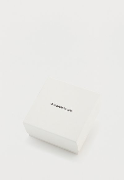 Boîte carrée blanche avec une texture lisse et un logo noir "Completedworks" imprimé sur le couvercle. Aucun autre motif ou élément métallique visible.