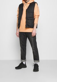 Svart vadderad väst över en orange hoodie, som bärs tillsammans med mörka jeans och svarta sneakers. Funktioner inkluderar fickor, en avslappnad passform och rullade ärmslut.