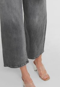 Henkilö, joka pukeutuu harmaisiin leveisiin farkkuihin ja läpinäkyviin korkokenkiin, seisoo vaaleanharmaalla lattialla.