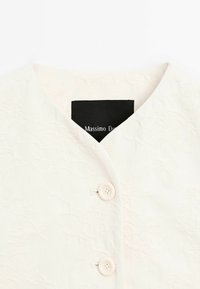 Vêtement blanc texturé à boutons avec deux boutons blancs et une étiquette rectangulaire noire partiellement visible à l'intérieur du col.