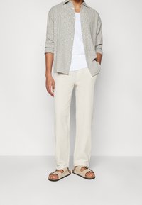 Een button-up overhemd in een zwart-wit ruitpatroon, gecombineerd met lichtbeige, textuurbroeken en bruine sandalen met banden.