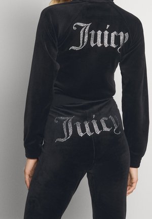 Czarny welurowy dres z logo „Juicy” z kryształków na górnej części pleców i na talii, noszony przez stojącą osobę zwróconą tyłem.