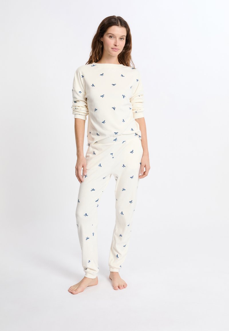 Conjunto de pijama en color crema que incluye una camiseta de manga larga y pantalones, adornados con pequeños motivos de lazo azul, hecho de una tela suave y con un corte relajado.