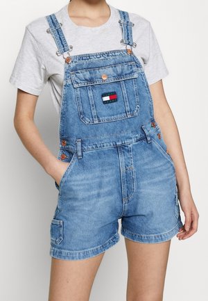 Personne portant une salopette courte en denim bleu clair avec poche avant et détails de boutons sur un t-shirt gris clair à manches courtes.