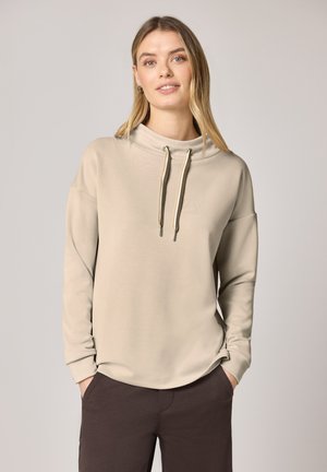 Młoda kobieta o długich blond włosach nosi beżową bluzę z kapturem z długim rękawem i sznurkami oraz ciemnobrązowe spodnie, ma ręce w kieszeniach, na tle o neutralnym kolorze.