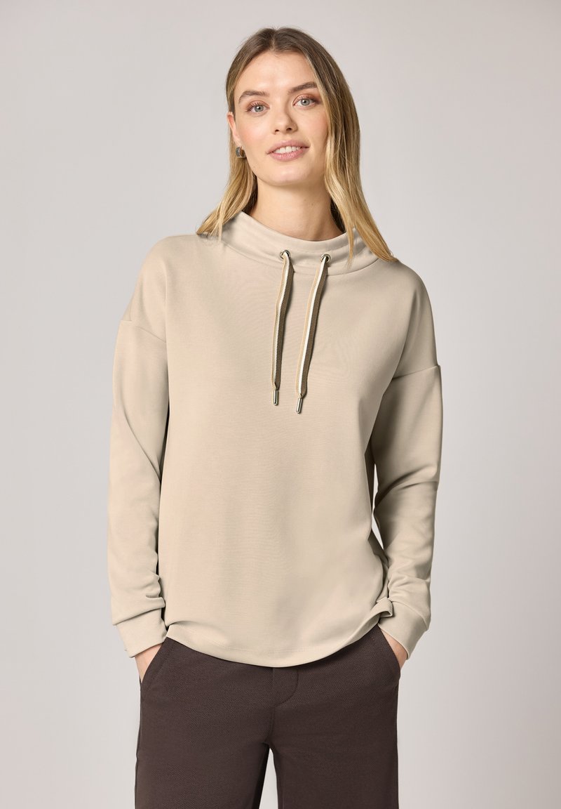 Mujer joven con cabello largo y rubio que lleva una sudadera beige de manga larga con cordones y pantalones marrón oscuro, con las manos en los bolsillos, fondo neutro.