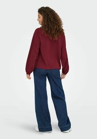 Bordeauxfarbene Bluse mit langen, ballonförmigen Ärmeln, kombiniert mit hoch taillierten, dunkelblauen Schlagjeans. Glatte Textur mit minimalen Akzentdetails.