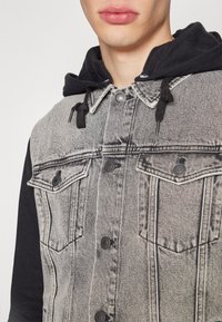 Närbild av en person som bär en grå acid-wash denimjacka med svarta knappar och en svart huva under med dragskor.
