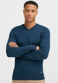 Tumesinine V-kaelusega sviiter sujuvast kudumisest, ribadega mansettide ja äärtega, väikese logo sildiga alumises vasakus nurgas.