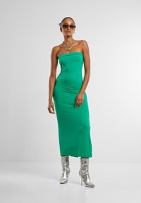 Žalia beplatė midi suknelė, pagaminta iš lygaus audinio, su bodycon siluetu. Su ja derinami blizgūs sidabriniai iki kelių batai ir saulės akiniai.