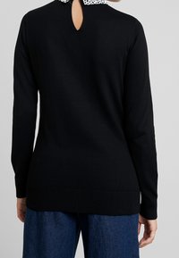 Svart stickad tröja med krage i prickar, långa ärmar och ett nyckelhål i ryggen. Har ribbat slut och muddar.