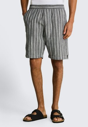 Szare męskie szorty z białymi pionowymi paskami, elastycznym pasem, bocznymi kieszeniami, noszone z czarnymi sandałami wsuwanymi na gołe nogi.