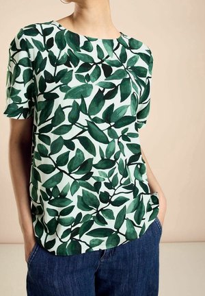 Vrouw draagt een wit T-shirt met korte mouwen met een donkergroen bladpatroon en blauwe spijkerbroek, handen in de zakken, beige achtergrond.