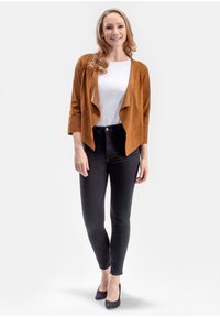 Bruine suède jas met vloeiende design, driekwartmouwen, gedragen over een witte T-shirt en zwarte skinny jeans, gecombineerd met zwarte hakken.