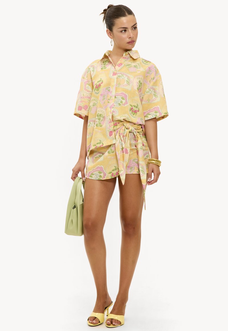 Ensemble chemise à manches courtes jaune vif avec motifs de fruits et short assorti ; accompagné d'un sac à main vert et de sandales à talons jaunes.