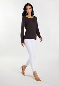 Braunes langärmliges Strickoberteil mit Rundhalsausschnitt, kombiniert mit enganliegenden weißen Jeans und nudefarbenen, spitz zulaufenden Absatzschuhen, die vor einem schlichten hellen Hintergrund laufen.