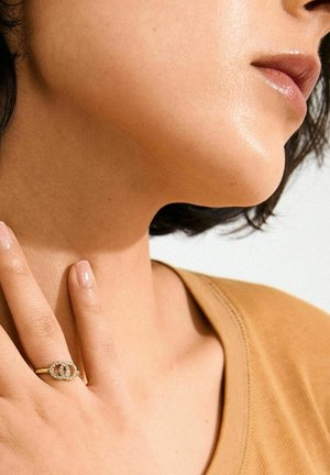 Bague en or avec deux cercles entrelacés, ornée de petites pierres claires. La main d'une personne touche doucement son cou. Fond neutre.