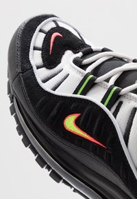 Černobílé atletické boty s textilním svrškem, texturovanými překryvy a neonovými akcenty v zelené a růžové barvě. Obsahují tkaničky a gumovou podrážku.