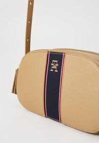 Borsa a tracolla in pelle lavorata color tan con una striscia blu navy e accenti rosso-bianco; logo con hardware dorato; chiusura con zip e tracolla regolabile.