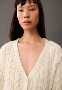 Cremefarvet kardigan i kabelstrik med dyb V-udskæring og en enkelt knaplukning. Tekstureret finish med fremtrædende snoede mønstre.