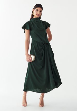 Vrouw draagt een donkergroene midijurk met zwierige mouwen, houdt een kleine gouden clutch vast en draagt gouden sandalen met bandjes en oorbellen.