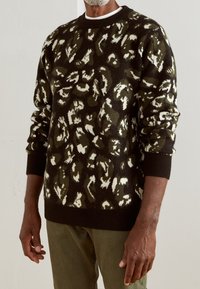 Pull en tricot avec une base noire présentant un motif abstrait vert et blanc. Poignets et col côtelés, tissu texturé, coupe décontractée.