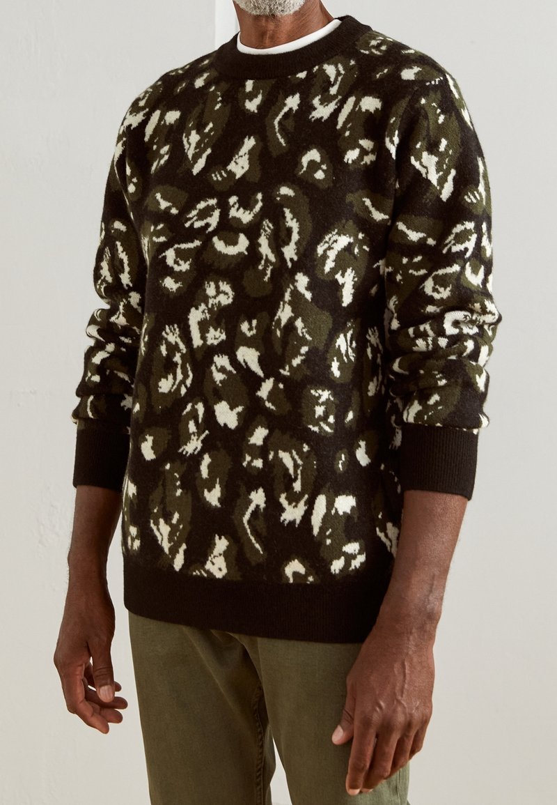 Pull en tricot avec une base noire présentant un motif abstrait vert et blanc. Poignets et col côtelés, tissu texturé, coupe décontractée.