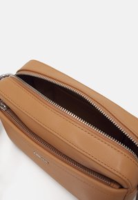 Sac bandoulière en cuir tan avec des finitions en métal argenté, comportant deux compartiments zippés et une texture lisse. L'intérieur présente une couleur contrastante.