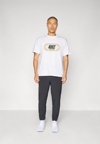 Biała koszulka Nike z graficznym logo, zestawiona z czarnymi spodniami i białymi trampkami. Koszulka ma luźny krój i miękką teksturę.
