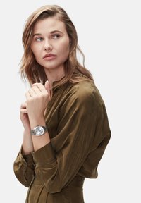 Silberne Uhr mit einem strukturierten runden Ziffernblatt, drei Unterzifferblättern und einem geflochtenen Metallarmband. Getragen an einem Handgelenk mit einem olivgrünen Satinhemd.