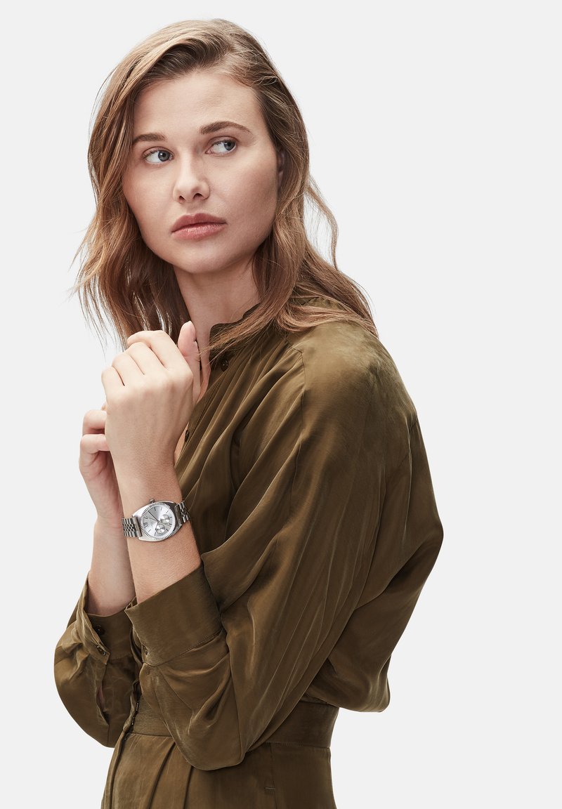 Silberne Uhr mit einem strukturierten runden Ziffernblatt, drei Unterzifferblättern und einem geflochtenen Metallarmband. Getragen an einem Handgelenk mit einem olivgrünen Satinhemd.
