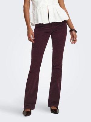 Femme portant un pantalon flare en velours côtelé violet foncé, un haut sans manches plissé blanc et des chaussures noires pointues, debout les mains détendues.
