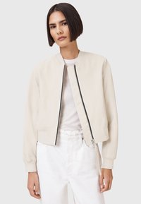 Chaqueta bomber de color beige claro con una textura suave, cierre delantero, puños acanalados y un diseño corto. Llevada sobre una camisa blanca y pantalones.