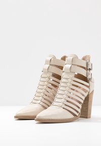 Chaussures à talons hauts beige avec un bout pointu, un design à lanières, un cuir texturé et un talon bloc en bois. Boucles en métal sur les brides de la cheville.