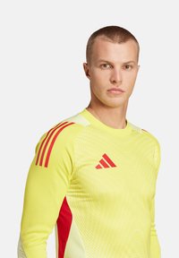 Heldere gele sportshirt met lange mouwen en rode accenten, voorzien van een gestructureerd patroon en drie rode strepen op elke schouder.