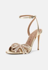 Goldene Sandale mit hohen Absätzen und Knöchelriemen, verziert mit einem mit Strass besetzten Schleifenmotiv auf der offenen Zehe, mit sichtbarem Guess-Innenfutter.
