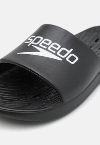 Crne sandale na klizanje s širokom trakom koja sadrži bijeli Speedo logo. Teksturirana unutrašnjost s drenažnim rupama za ventilaciju. Izdržljiv materijal.