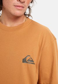 Brown cotton t-shirt featuring a black wave logo on the left chest. Short sleeves and round neckline with a smooth texture.