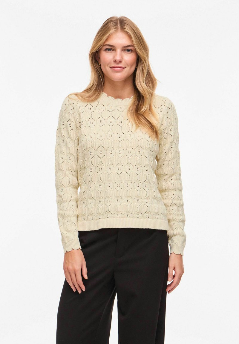 Femme blonde aux cheveux mi-longs portant un pull en maille crème texturé et un pantalon noir, debout devant un fond blanc uni.
