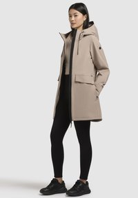 Beige vandtæt jakke med hætte, frontlynlås og to store lommer. Har sorte accenter og et slankt, moderne design.