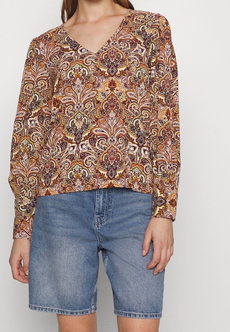 Femme portant un chemisier à manches longues col en V avec un motif paisley complexe orange et marron et un short en jean bleu mi-long.